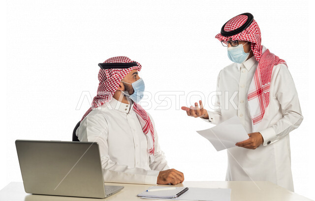 بورتريه لموظفان خليجيان سعوديان في مقر العمل، إرتداء القناع الطبي الوقائي للوقاية من فايروس كورونا كوفيد-19، إستخدام الحاسوب المحمول لإنجاز المهام، العمل عن بعد، زملاء عمل، شركة سعودية،أعمال تجارية، مفهوم إدارة الأعمال، مناقشة العمل، مراجعة البحوث العلمية