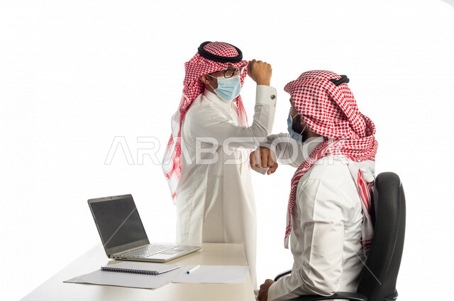 بورتريه لموظفان خليجيان سعوديان في مقر العمل يتصافحان عن بعد، إرتداء القناع الطبي الوقائي للوقاية من فايروس كورونا كوفيد-19، إستخدام الحاسوب المحمول لإنجاز المهام، زملاء عمل، مفهوم إدارة الأعمال، إتباع إجراءات الوقاية من الفيروسات، الحفاظ على المسافات