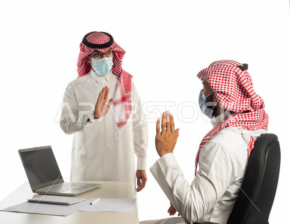 بورتريه لموظفان خليجيان سعوديان في مقر العمل يتصافحان عن بعد، إرتداء القناع الطبي الوقائي للوقاية من فايروس كورونا كوفيد-19، إستخدام الحاسوب المحمول لإنجاز المهام، زملاء عمل، مفهوم إدارة الأعمال، إتباع إجراءات الوقاية من الفيروسات، الحفاظ على المسافات