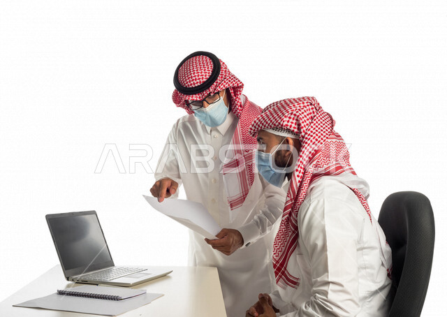 بورتريه لموظفان خليجيان سعوديان في مقر العمل، إرتداء القناع الطبي الوقائي للوقاية من فايروس كورونا كوفيد-19، إستخدام الحاسوب المحمول لإنجاز المهام، العمل عن بعد، زملاء عمل، شركة سعودية،أعمال تجارية، مفهوم إدارة الأعمال، مناقشة العمل، مراجعة البحوث العلمية