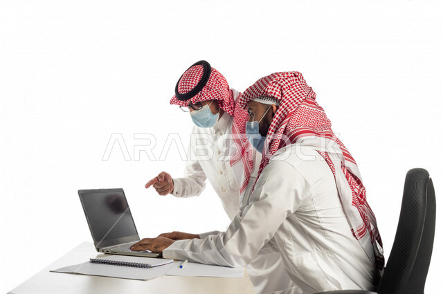 بورتريه لموظفان خليجيان سعوديان في مقر العمل، إرتداء القناع الطبي الوقائي للوقاية من فايروس كورونا كوفيد-19، إستخدام الحاسوب المحمول لإنجاز المهام، العمل عن بعد، زملاء عمل، شركة سعودية،أعمال تجارية، مفهوم إدارة الأعمال، مناقشة العمل، مراجعة البحوث العلمية
