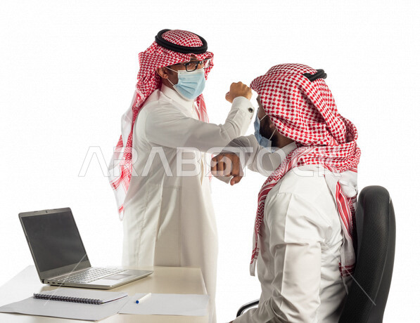 بورتريه لموظفان خليجيان سعوديان في مقر العمل يتصافحان عن بعد، إرتداء القناع الطبي الوقائي للوقاية من فايروس كورونا كوفيد-19، إستخدام الحاسوب المحمول لإنجاز المهام، زملاء عمل، مفهوم إدارة الأعمال، إتباع إجراءات الوقاية من الفيروسات، الحفاظ على المسافات