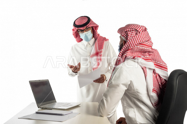 بورتريه لموظفان خليجيان سعوديان في مقر العمل، إرتداء القناع الطبي الوقائي للوقاية من فايروس كورونا كوفيد-19، إستخدام الحاسوب المحمول لإنجاز المهام، العمل عن بعد، زملاء عمل، شركة سعودية،أعمال تجارية، إتباع إجراءات الوقاية، مفهوم إدارة الأعمال، مناقشة العمل