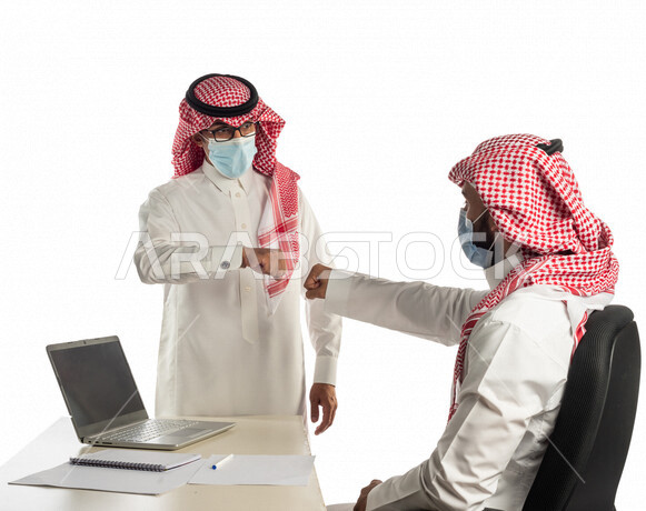 بورتريه لموظفان خليجيان سعوديان في مقر العمل يتصافحان عن بعد، إرتداء القناع الطبي الوقائي للوقاية من فايروس كورونا كوفيد-19، إستخدام الحاسوب المحمول لإنجاز المهام، زملاء عمل، مفهوم إدارة الأعمال، إتباع إجراءات الوقاية من الفيروسات، الحفاظ على المسافات