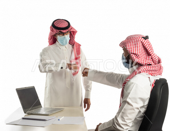 بورتريه لموظفان خليجيان سعوديان في مقر العمل يتصافحان عن بعد، إرتداء القناع الطبي الوقائي للوقاية من فايروس كورونا كوفيد-19، إستخدام الحاسوب المحمول لإنجاز المهام، زملاء عمل، مفهوم إدارة الأعمال، إتباع إجراءات الوقاية من الفيروسات، الحفاظ على المسافات