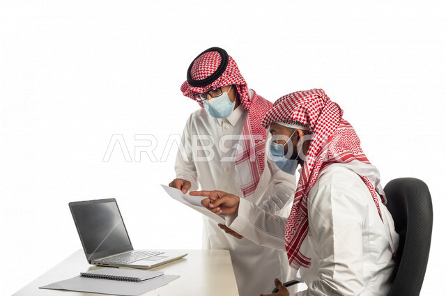 بورتريه لموظفان خليجيان سعوديان في مقر العمل، إرتداء القناع الطبي الوقائي للوقاية من فايروس كورونا كوفيد-19، إستخدام الحاسوب المحمول لإنجاز المهام، العمل عن بعد، زملاء عمل، شركة سعودية،أعمال تجارية، مفهوم إدارة الأعمال، مناقشة العمل، مراجعة البحوث العلمية