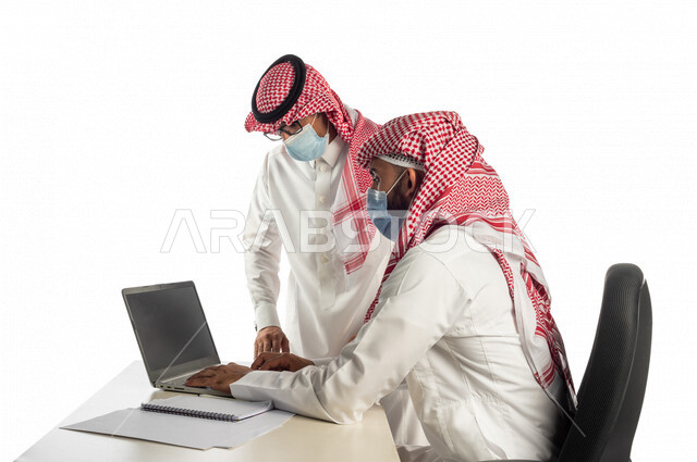 بورتريه لموظفان خليجيان سعوديان في مقر العمل، إرتداء القناع الطبي الوقائي للوقاية من فايروس كورونا كوفيد-19، إستخدام الحاسوب المحمول لإنجاز المهام، العمل عن بعد، زملاء عمل، شركة سعودية،أعمال تجارية، مفهوم إدارة الأعمال، مناقشة العمل، مراجعة البحوث العلمية