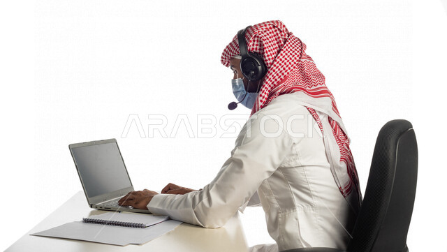 بورتريه لرجل أعمال خليجي سعودي يعمل في خدمة العملاء، مرتدي القناع الطبي للوقاية من فايروس كورونا كوفيد-19، يضع سماعة الاتصال، استعلامات واستفسارات، اجراء اتصال فيديو من خلال اللابتوب، العمل عن بعد، عقد اجتماع عمل عبر الانترنت، جلسة عمل، التسويق الإلكتروني