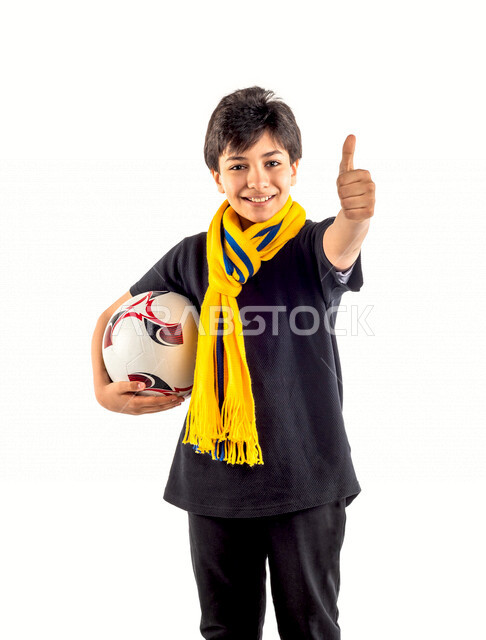 بورتريه لطفل عربي خليجي سعودي يحمل كرة قدم تحت ذراعه، يرتدي وشاح فريق النصر الرياضي، إيماءات وجه سعيدة ويدين تدل على الموافقة والاعجاب، خلفية بيضاء