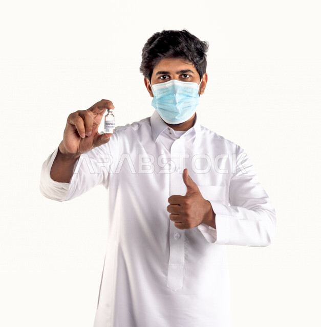 Portrait of a Saudi Arabian Gulf youth wearing a mask and holding the vaccine package against Corona Virus (Covid-19) in his hand, pointing with his thumb finger up, vaccinations against diseases, with facial gestures indicating acceptance, approval and a