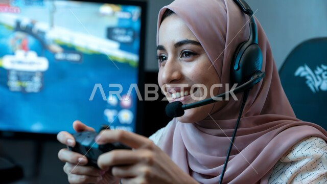 الاستمتاع بالتقنيات والتكنولوجيا الحديثة، مفهوم المتعة والتسلية باللعب، صورة مقربة لامرأة عربية سعودية خليجية محجبة مبتسمة ترتدي سماعة الرأس تمسك بيدها عصا التحكم تلعب بألعاب الفيديو بإيماءات السعادة والفرح، برامج الكمبيوتر الترفيهية