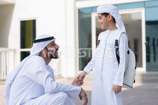 التحفيز المعنوي للأبناء في أول أيام الدوام المدرسي، إلقاء عبارات التشجيع الداعمة وتقديم الدعم النفسي، موسم العودة إلى المدارس بالامارات العربية المتحدة، مفهوم الأبوة والحنان، ولد عربي خليجي إماراتي يرتدي الكندورة والعمامة حقيبة الظهر المدرسية ويقف برفقة والده