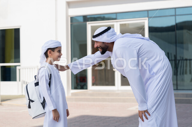 تقديم الدعم النفسي والتحفيز المعنوي للأبناء في أول أيام الدوام المدرسي، إلقاء عبارات التشجيع الداعمة، موسم العودة إلى المدارس بالامارات العربية المتحدة، مفهوم الأبوة والحنان، ولد عربي خليجي إماراتي يرتدي الكندورة والعمامة وحقيبة الظهر المدرسية يقف برفقة والده