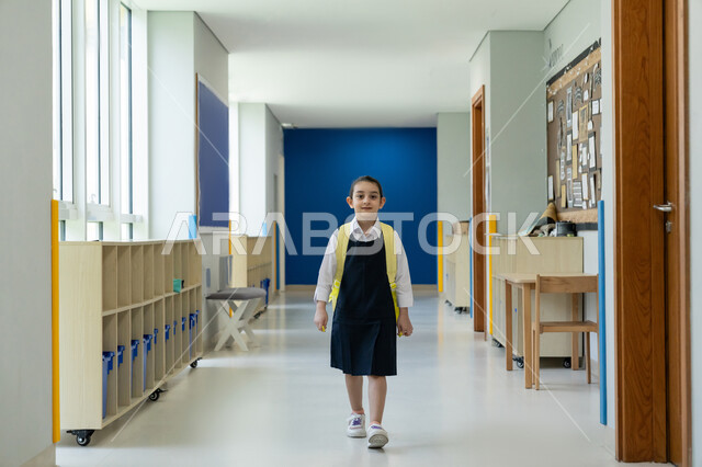 التفاؤل والحماس في موسم العودة للمدارس, التعرف على الفصول الدراسية وتفاصيل المدرسة, التعليم الأكاديمي في المؤسسات التعليمية بالإمارات العربية المتحدة, طالبة عربية خليجية إماراتية ترتدي الزي المدرسي وتحمل حقيبة الظهر تقف في ممر المدرسة في أول يوم دراسي لها