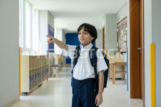 الوقوف في ممر المدرسة بسعادة وتفاؤل، المؤسسات التعليمية المتطورة في الامارات العربية المتحدة، البدء بعام دراسي جديد بإيماءات الحماس، السعي نحو تحقيق التفوق والنجاح، صورة مقربة لتلميذ عربي خليجي إماراتي يرتدي الزي المدرسي ويحمل حقيبة الظهر  يشير إلى مكان ما