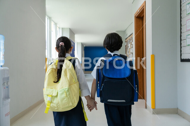 السعي للتفوق وتحقيق النجاح، الانطلاق والتوجه نحو الفصل الدراسي المخصص، تقوية علاقة وروابط المحبة عبر إمساك الأخوة لأيدي بعضهم البعض، صورة مقربة من الخلف لأخ وأخته عربيين خليجيين إماراتيين يرتديان الزي المدرسي الرسمي ويحملان حقائب الظهر يقفان في ممر المدرسة