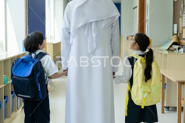 الإمساك بأيدي الأبناء لتعزيز مفهوم الأمان لديهم، تحفيز الأولاد وتشجيعهم في بداية السنة الدراسية، صورة مقربة من الخلف لأب عربي خليجي إماراتي يرتدي الكندورة والغترة يرافق ابناءه إلى المدرسة في يومهم الأول، أخ واخته يحملان حقائب الظهر ويقفان في الممر المدرسي