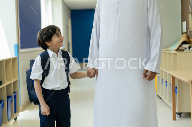 تعزيز مفهوم الأمان لدى الأبناء، تشجيع الأولاد وتحفيزهم في بداية السنة الدراسية، السعادة والحماس بالعودة إلى المدرسة، صورة مقربة لأب عربي خليجي إماراتي يرتدي الكندورة والغترة يرافق ابنه إلى المدرسة في يومه الدراسي الأول، تلميذ مرتب يحمل حقيبة الظهر بفرح