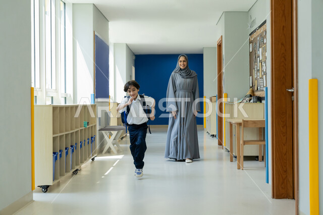 Expressions of happiness and enthusiasm for the start of the new school year, early attendance at school classes, interest in educating and developing children and supporting them, a veiled Emirati Gulf Arab mother wearing a gray abaya accompanies her son on his first day of school, an Emirati student wearing a school uniform and carrying a backpack with joy