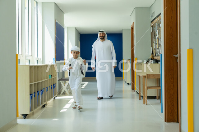 توديع ومرافقة الأبناء إلى الصف في أول أيام الفصل الدراسي، إلقاء عبارات التشجيع الداعمة وتقديم الدعم النفسي، موسم العودة إلى المدارس بالامارات العربية المتحدة، ولد عربي خليجي إماراتي يرتدي الكندورة والعمامة وحقيبة الظهر المدرسية برفقة والده يجري في ممر المدرسة