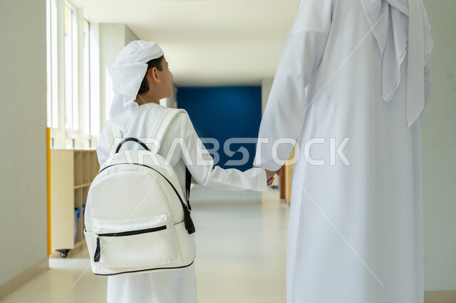 Back to school season in the UAE, strengthening the parental bond and providing moral and psychological support to children, accompanying children to class on the first day of the school term, close-up from the back of an Emirati Gulf Arab boy wearing a kandura, turban and backpack and holding his father's hand