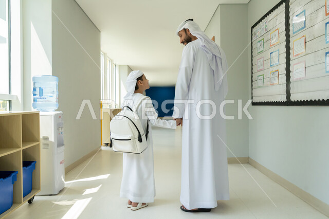 تقوية رابط الأبوة وتقديم الدعم المعنوي والنفسي للأطفال، توديع ومرافقة الأبناء إلى الصف في أول أيام الفصل الدراسي، موسم العودة إلى المدارس بالامارات العربية المتحدة، صورة من الخلف لولد عربي خليجي إماراتي يرتدي الكندورة والعمامة وحقيبة الظهر ويمسك بيد والده