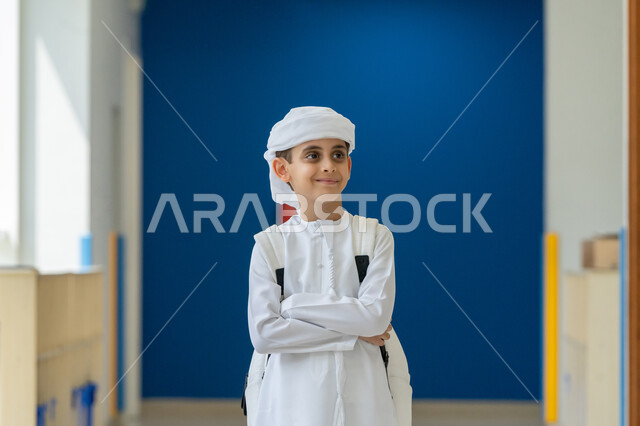 النظر الى شيء ما بتعابير السعادة والسرور، مفهوم الاهتمام بالمظهر الخارجي, طالب عربي خليجي إماراتي يرتدي الكندورة والعمامة يحمل الحقيبة المدرسية ويقف باستقامة بأيدي متقاطعة، موسم العودة الى المدارس، ايماءات الفرح بالنجاح وتحقيق الأهداف