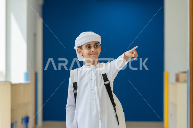 Specifying a specific place, the development and progress of educational curricula in the Kingdom of Saudi Arabia, gestures of joy at success and achieving goals, an Arab Gulf Emirati student wearing a kandura and turban carrying a school bag and pointing with his index finger at something