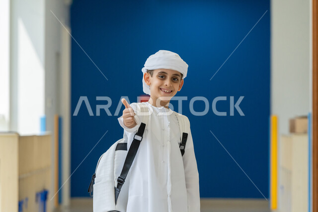 The concept of back to school, seriousness and diligence to achieve success, an Arab Gulf Emirati student wearing a kandura and turban carrying a school bag and raising his thumb with gestures of admiration and satisfaction, educational scientific curricula, looking at the camera with expressions of approval and happiness