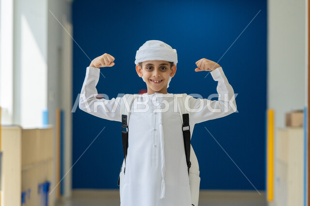 الانتصار والفوز والنجاح، مفهوم الانجاز وتحقيق الاهداف، طالب عربي خليجي إماراتي يرتدي الكندورة والعمامة يحمل الحقيبة المدرسية يرفع يديه للاعلى بإيماءات الفرح والسرور, الشعور بالسعادة والقوة, النظر الى الكاميرا بتعابير الفخر