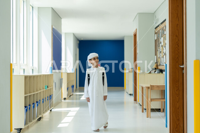 الجد والاجتهاد للوصول إلى النجاح، طالب عربي خليجي إماراتي يرتدي الكندورة والعمامة يحمل الحقيبة المدرسية ويمشي في أحد ممرات المدرسة، مفهوم العودة إلى المدارس، تطور وتقدم المناهج التعليمية في الامارات العربية المتحدة، إيماءات الفرح بالنجاح وتحقيق الأهداف