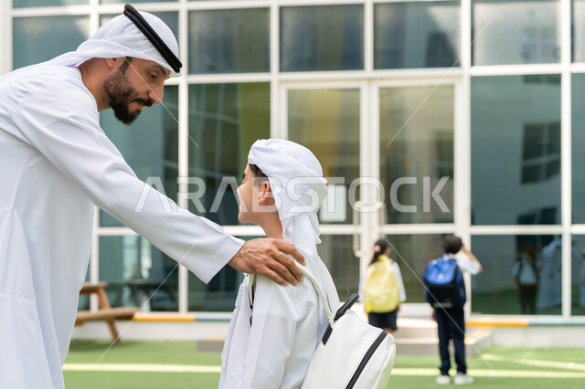 Giving psychological support and moral motivation to children, back-to-school season in the United Arab Emirates, the concept of fatherhood and tenderness, an Arab Gulf Emirati boy wearing kandora and ghutra and wearing a school backpack with his father, giving supportive words of encouragement on the first day of school