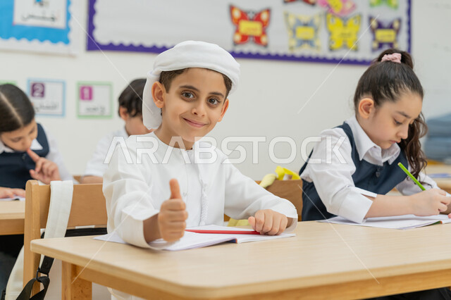 Education goals in Saudi Arabia, Arab Gulf Emirati students sitting on desks in the classroom and writing their homework, a diligent student raising his thumb in appreciation and satisfaction, developing creative skills, modern learning methods