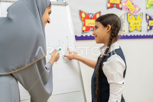 Improving the educational level in schools, a Saudi Arabian Gulf teacher wearing an abaya helps a student write Arabic letters on the board, interaction and activity in the classroom, interest in science and education in the United Arab Emirates