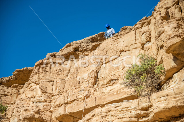 صورة لشاب عربي خليجي سعودي، ممارسة رياضة الهايكينج، تسلق الجبال الصخرية، حجارة وصخور جبلية، صحة ولياقة بدنية، استخدام الحبال في تسلق الجبال