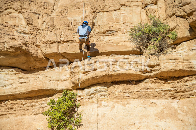 صورة لشاب عربي خليجي سعودي، ممارسة رياضة الهايكينج، تسلق الجبال الصخرية، حجارة وصخور جبلية، صحة ولياقة بدنية، استخدام الحبال في تسلق الجبال