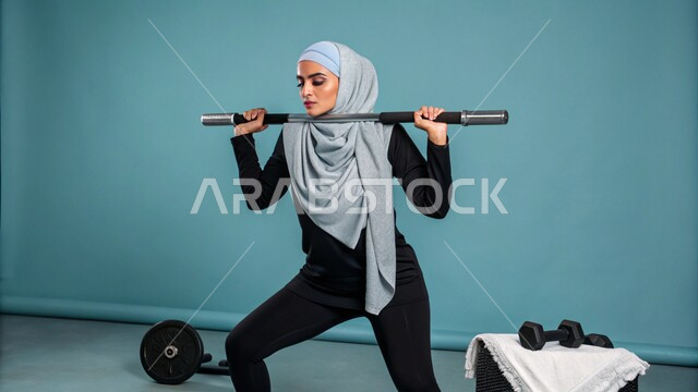 ممارسة التمارين الرياضيه و رفع الأثقال (دمبلز), المحافظة على الصحه واللياقه الجسديه و اللياقة الجسدية، حرق الدهون والسعرات الحراريه, بورتريه لإمرأه عربيه سعوديه خليجيه محجبه ترتدي لباس الرياضه تحمل بيديها الاوزان الحديدية الثقيلة، خلفية زرقاء