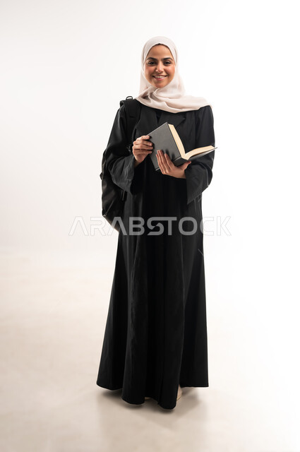 المناهج والمواد التعليمية الحديثة، التعليم العالي في جامعات المملكة، الاستعداد للعام الجامعي الجديد، بورتريه لطالبة جامعية عربية خليجية سعودية محجبة ترتدي حقيبة الظهر تقوم بتصفح كتاب بايماءات السرور، صورة لكامل الجسم، خلفية بيضاء
