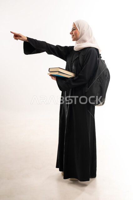 المناهج والمواد التعليمية الحديثة، التعليم العالي في جامعات المملكة، الاستعداد للعام الجامعي الجديد، بورتريه من الجانب لطالبة جامعية عربية خليجية سعودية محجبة ترتدي حقيبة الظهر تحمل مجموعة من الكتب تشير باصبع السبابة لمكان ما، صورة لكامل الجسم، خلفية بيضاء