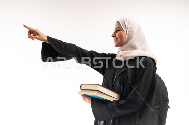Higher education in the universities of the Kingdom, modern curricula and educational materials, preparing for the new academic year, close-up portrait from the side of a veiled Saudi Arabian Gulf Arab female university student wearing a backpack carrying a set of books pointing with her index finger to a place, white background