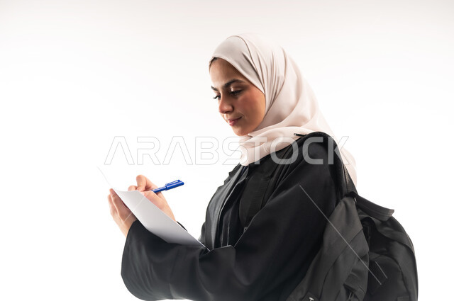 الاستعداد للعام الجامعي الجديد، الجامعات والكليات الأكاديمية، بورتريه مقرب من الجانب لطالبة جامعية عربية خليجية سعودية محجبة ترتدي عباءة سوداء تحمل حقيبة الظهر تمسك بيدها ورقة وقلم تقوم بمتابعة الدروس اليومية، خلفية بيضاء