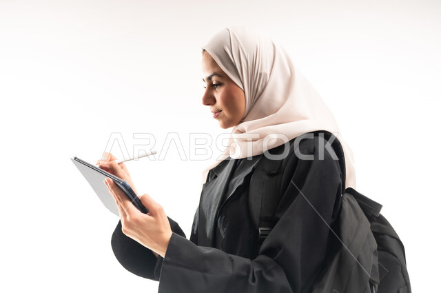 المواد والمناهج التعليمية في المملكة، مفهوم التعليم عن بعد، متابعة المحاضرات عبر المنصات التعليميه المتطوره، بورتريه مقرب من الجانب لطالبة جامعية عربية خليجية سعودية محجبة ترتدي عباءة سوداء تحمل حقيبة الظهر وجهاز التابلت، خلفية بيضاء