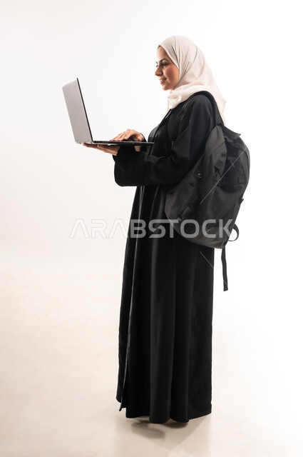 المناهج والمواد التعليمية في المملكة، مفهوم التعليم عن بعد، استخدام الأجهزة التقنية الحديثة لمتابعة المحاضرات، بورتريه من الجانب لطالبة جامعية عربية خليجية سعودية محجبة ترتدي عباءة سوداء تحمل حقيبة الظهر والحاسوب المحمول، صورة لكامل الجسم، خلفية بيضاء