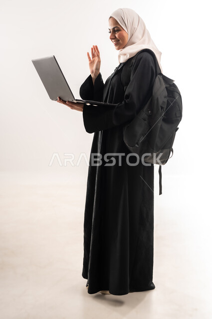 استخدام الأجهزة التقنية الحديثة لمتابعة المحاضرات، مفهوم التعليم عن بعد، المناهج والمواد التعليمية في المملكة، بورتريه مقرب من الجانب لطالبة جامعية عربية خليجية سعودية محجبة تحمل حقيبة الظهر والحاسوب المحمول ترفع يدها بايماءات التفاعل، صورة لكامل الجسم، خلفية بيضاء