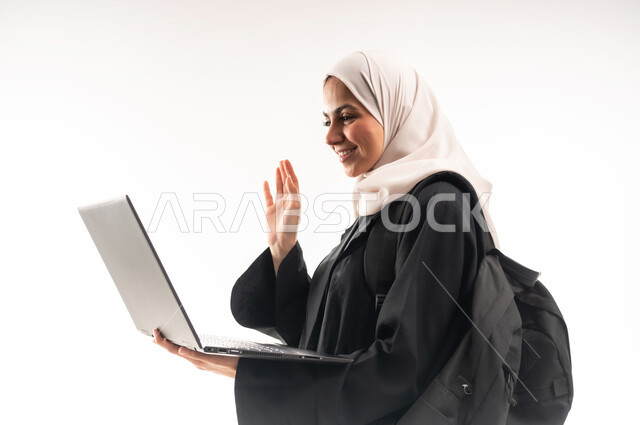 مفهوم التعليم عن بعد، المناهج والمواد التعليمية في المملكة، استخدام الأجهزة التقنية الحديثة لمتابعة المحاضرات، بورتريه مقرب من الجانب لطالبة جامعية عربية خليجية سعودية محجبة تحمل حقيبة الظهر والحاسوب المحمول ترفع يدها بايماءات التفاعل، خلفية بيضاء