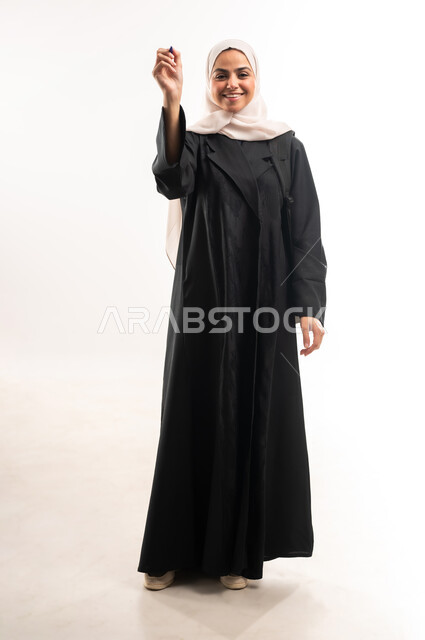 Preparing to write down and outline something, concept of drawing and planning, portrait of a veiled Saudi Arabian Gulf university student carrying a backpack holding a pen and making writing gestures in the air, full body image, white background