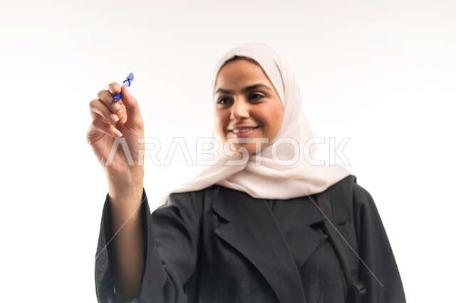 Concept of drawing and sketching, getting ready to write down and outline something, close-up portrait of a veiled Saudi Arabian Gulf university student carrying a backpack holding a pen and making writing gestures in the air, white background