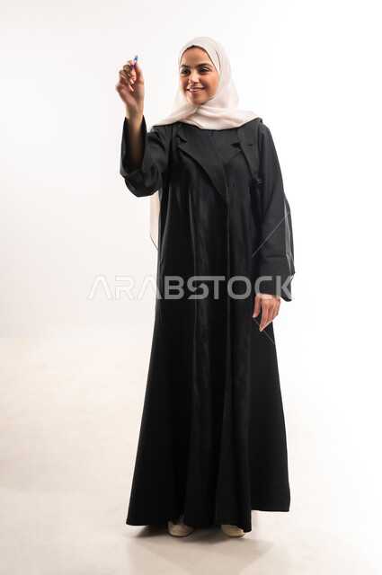 Preparing to write down and outline something, concept of drawing and planning, portrait of a veiled Saudi Arabian Gulf university student carrying a backpack holding a pen and making writing gestures in the air, full body image, white background