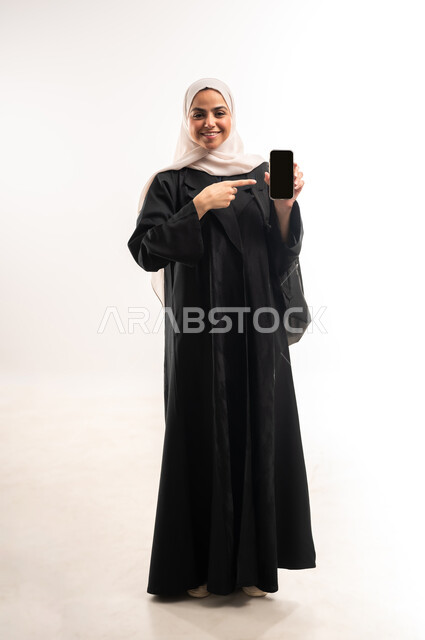 Looking at camera with happy gestures, showing blank black screen, using modern technology devices in daily life, university education in the Kingdom, portrait of a veiled Saudi Arabian Gulf female university student holding a backpack pointing to a mobile phone, full body photo, white background