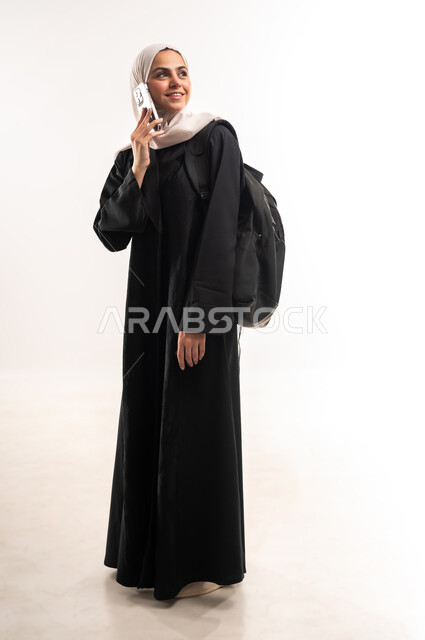 إجراء اتصال هاتفي عبر الجوال, استخدام الأجهزة التقنية الحديثة في الحياة اليومية، التعليم الجامعي في المملكة، بورتريه لطالبة جامعية عربية خليجية سعودية محجبة ترتدي عباءة سوداء تحمل حقيبة الظهر تمسك بيدها الهاتف المحمول، صورة لكامل الجسم، خلفية بيضاء